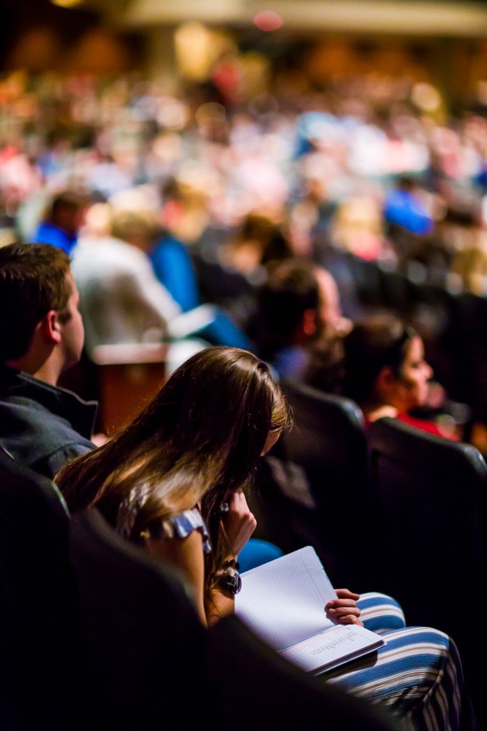 A focused shot of a person reading amidst a large audience in an indoor auditorium.